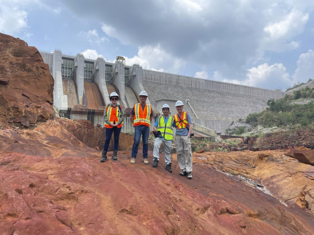 Four individuals wearing hard hats and high-visibility clothing stand on rocky terrain in front of a large dam structure under a cloudy sky.
