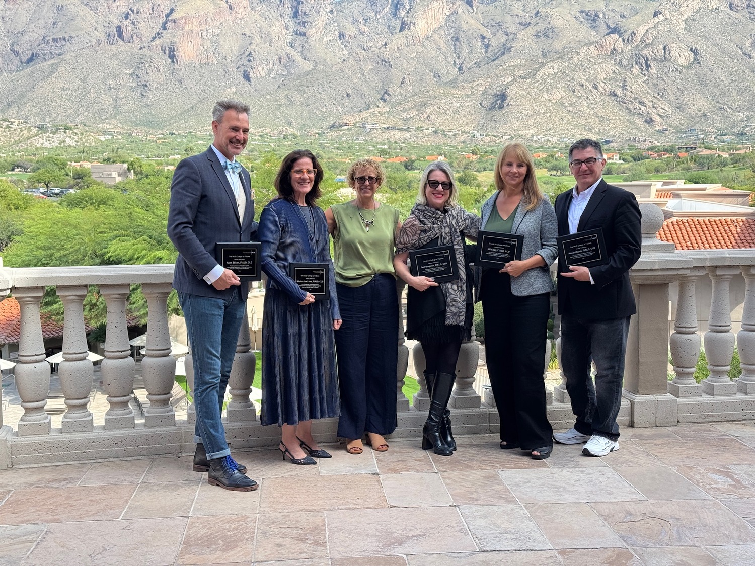 Six people standing on a terrace with a mountain view; five hold plaques, suggesting an award ceremony or recognition event.