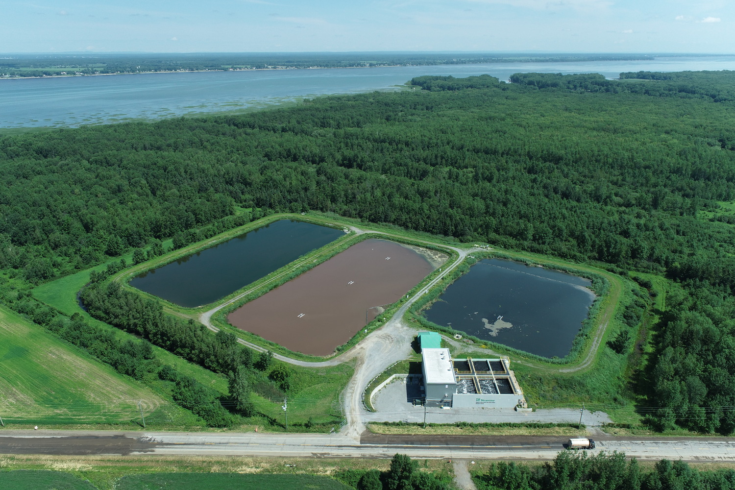 Aerial view of a water treatment facility with two large settling ponds and a main processing building, surrounded by dense forest and located near a body of water.
