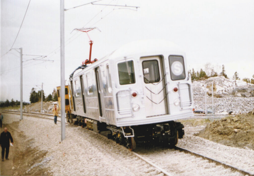 A silver subway train with an "S" sign is being tested on an outdoor track, with overhead wires and a construction site in the background.