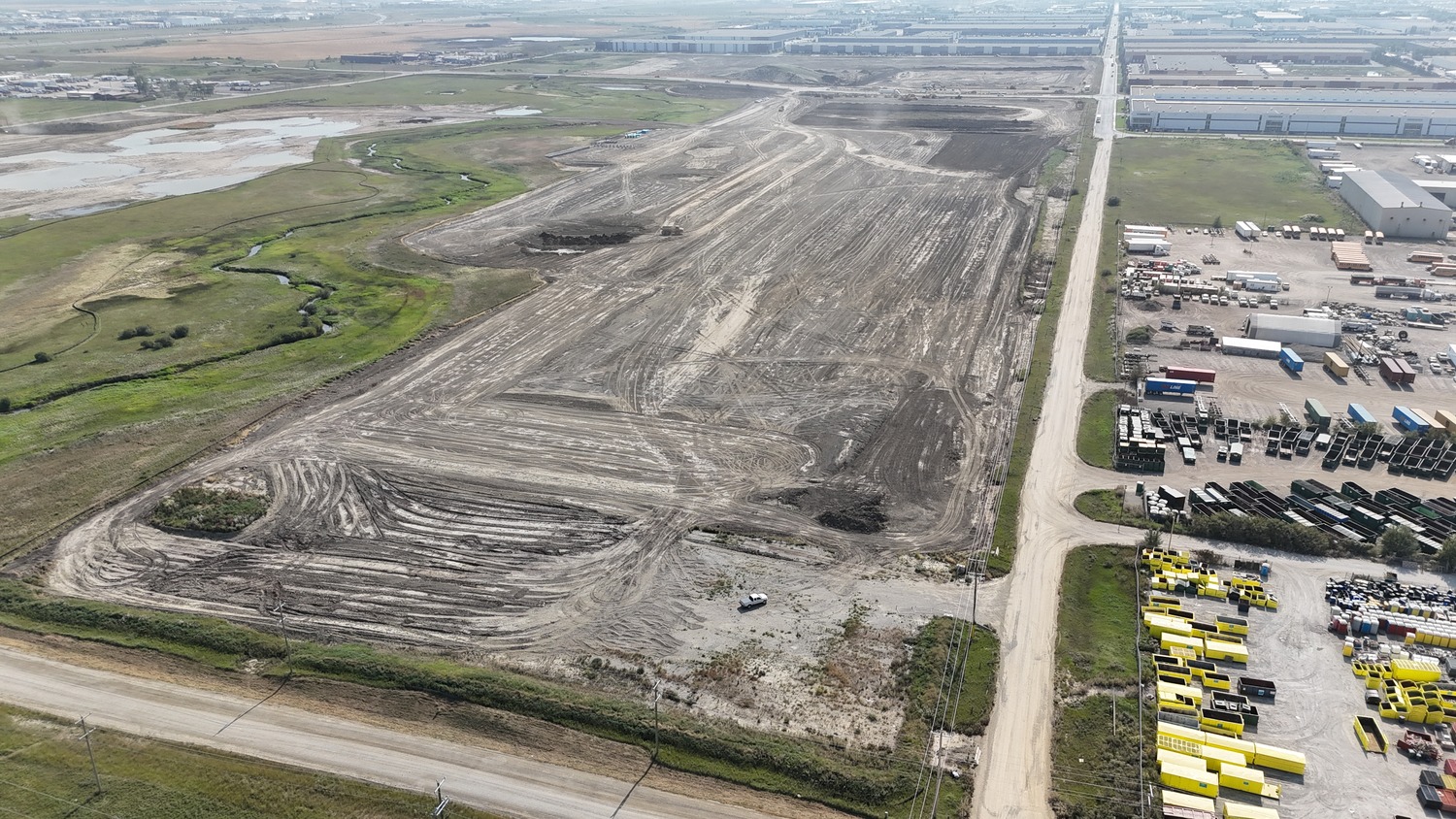 Aerial view of a large construction site with cleared land, dirt roads, machinery, and adjacent industrial buildings and equipment yards.