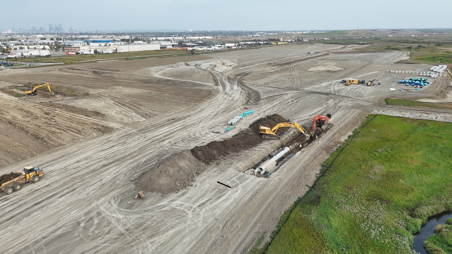 Aerial view of a construction site with excavators digging and moving earth, large pipes being laid, and machinery scattered across a wide, cleared area near a grassy patch.
