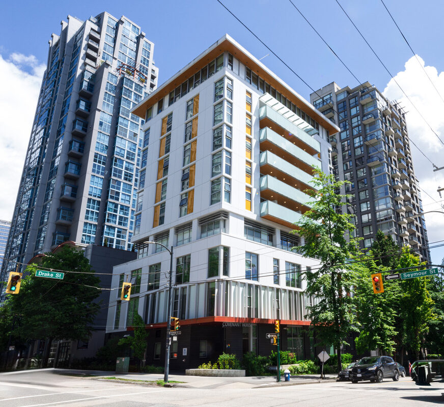 Modern multi-story residential and commercial building at a city intersection, surrounded by tall glass and steel high-rises under a clear blue sky.
