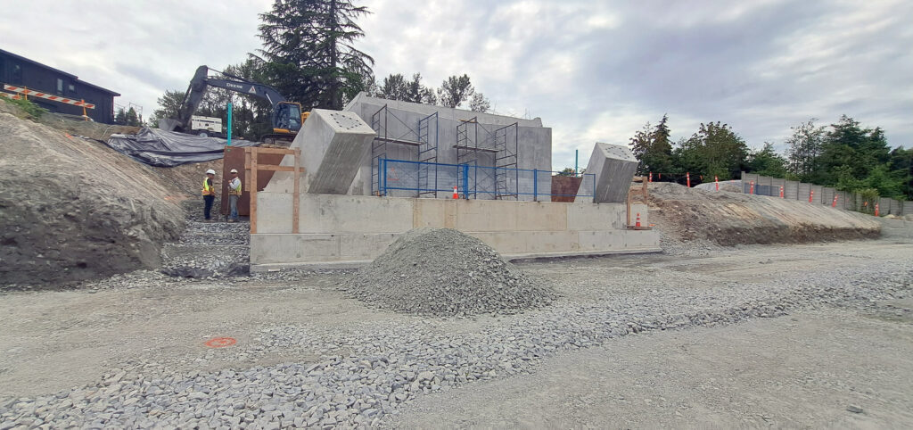 Construction site with concrete walls being built, scaffolding, gravel piles, a worker in a safety vest, and an excavator in the background, surrounded by trees and fencing.