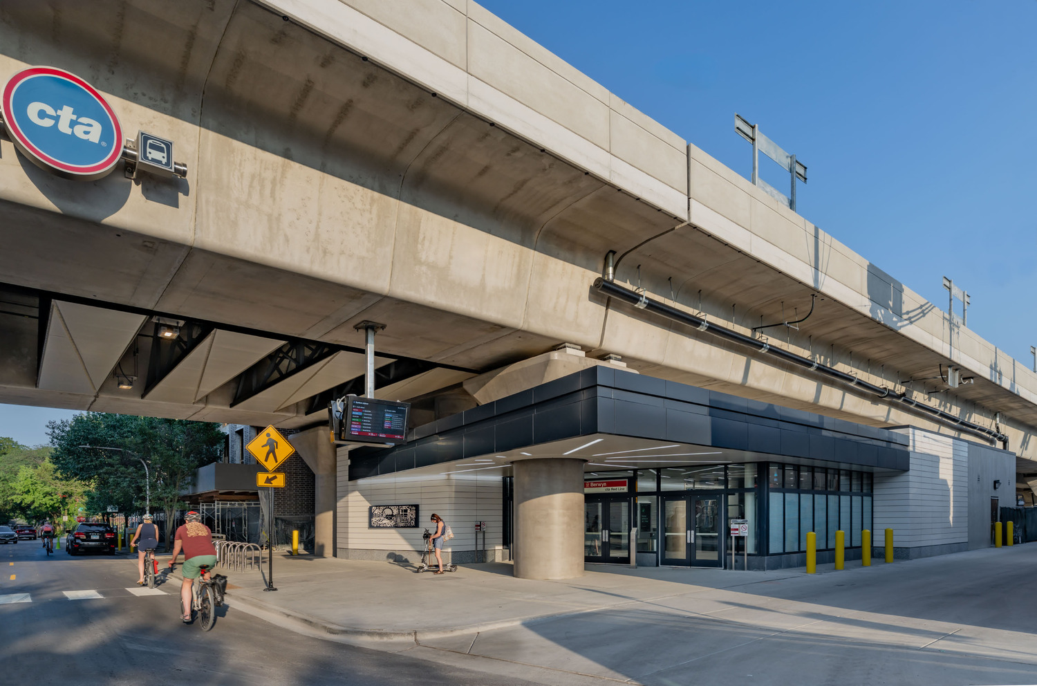 A modern CTA transit station entrance is located beneath an elevated railway track, with people walking, biking, and cars exiting the area.