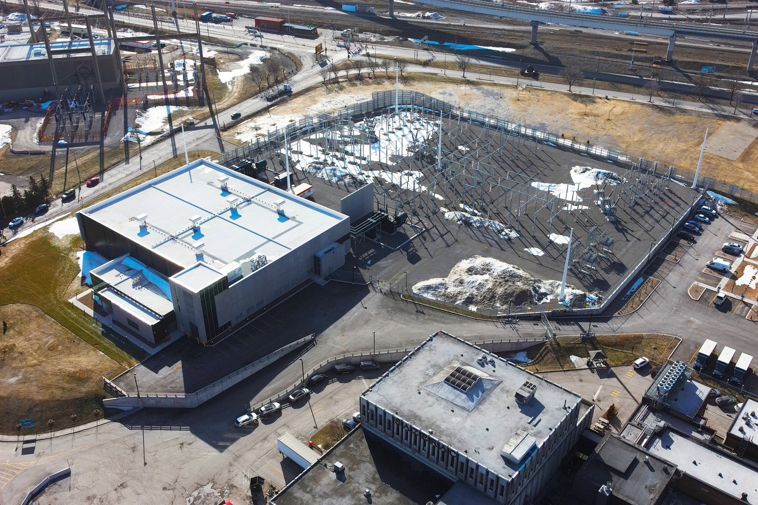 Aerial view of an industrial facility with a main building, adjacent fenced area with metal structures, surrounding roads, and patches of snow on the ground.