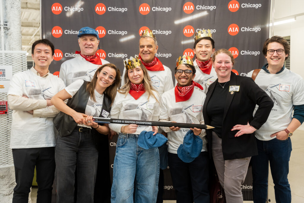 A group of nine people wearing matching shirts and red bandanas pose indoors with a sign. The background displays a board with "AIA Chicago." Two wear crowns, and most are smiling.