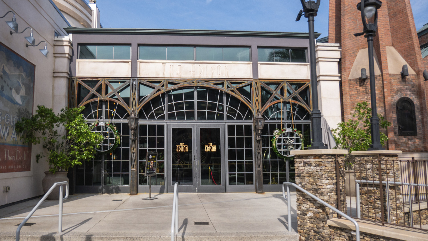 Exterior of a brick and glass building with double doors, featuring decorative metalwork and surrounded by stone planters and outdoor lights.
