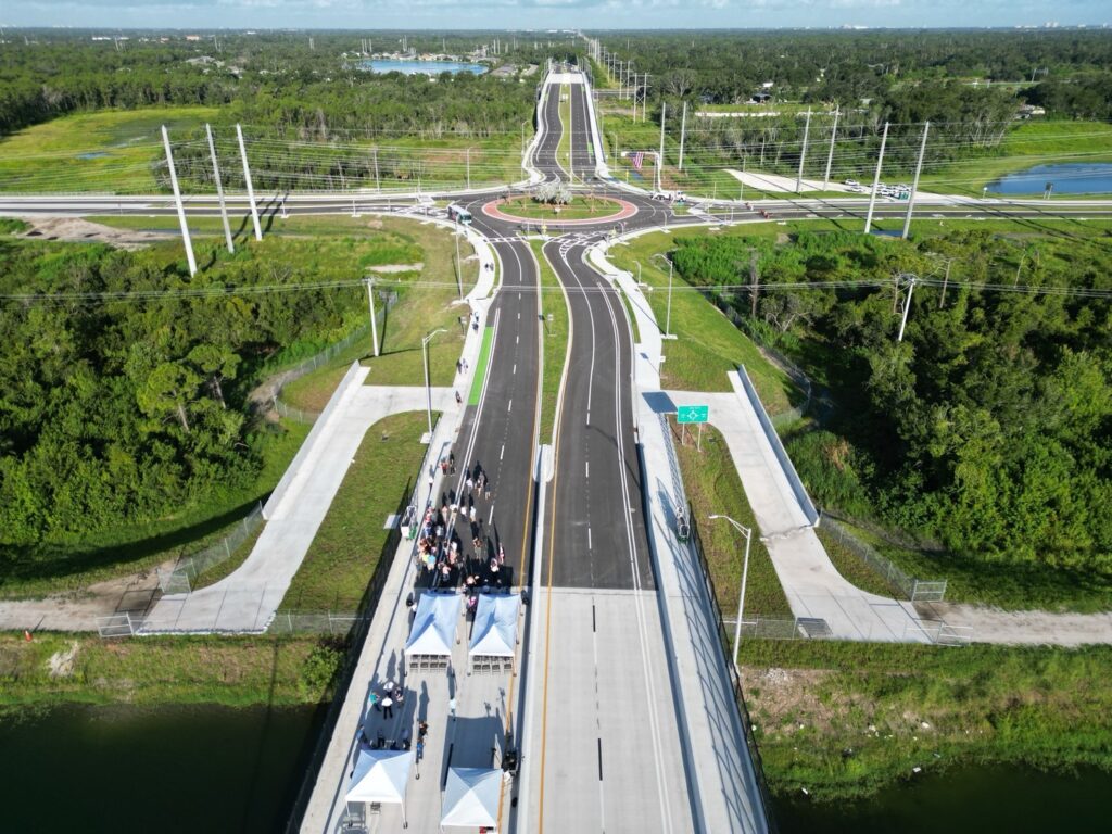 Aerial view of a newly constructed road and bridge with people gathered under white tents for an event near the bridge entrance, surrounded by green landscape and power lines.