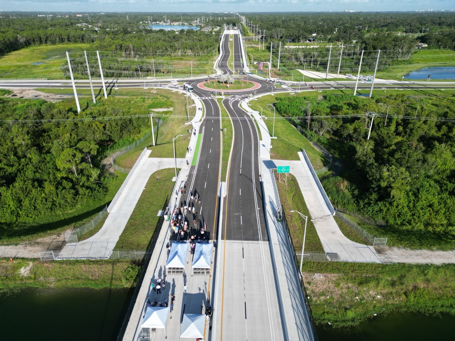 Aerial view of a newly constructed road and bridge with people gathered under white tents for an event near the bridge entrance, surrounded by green landscape and power lines.