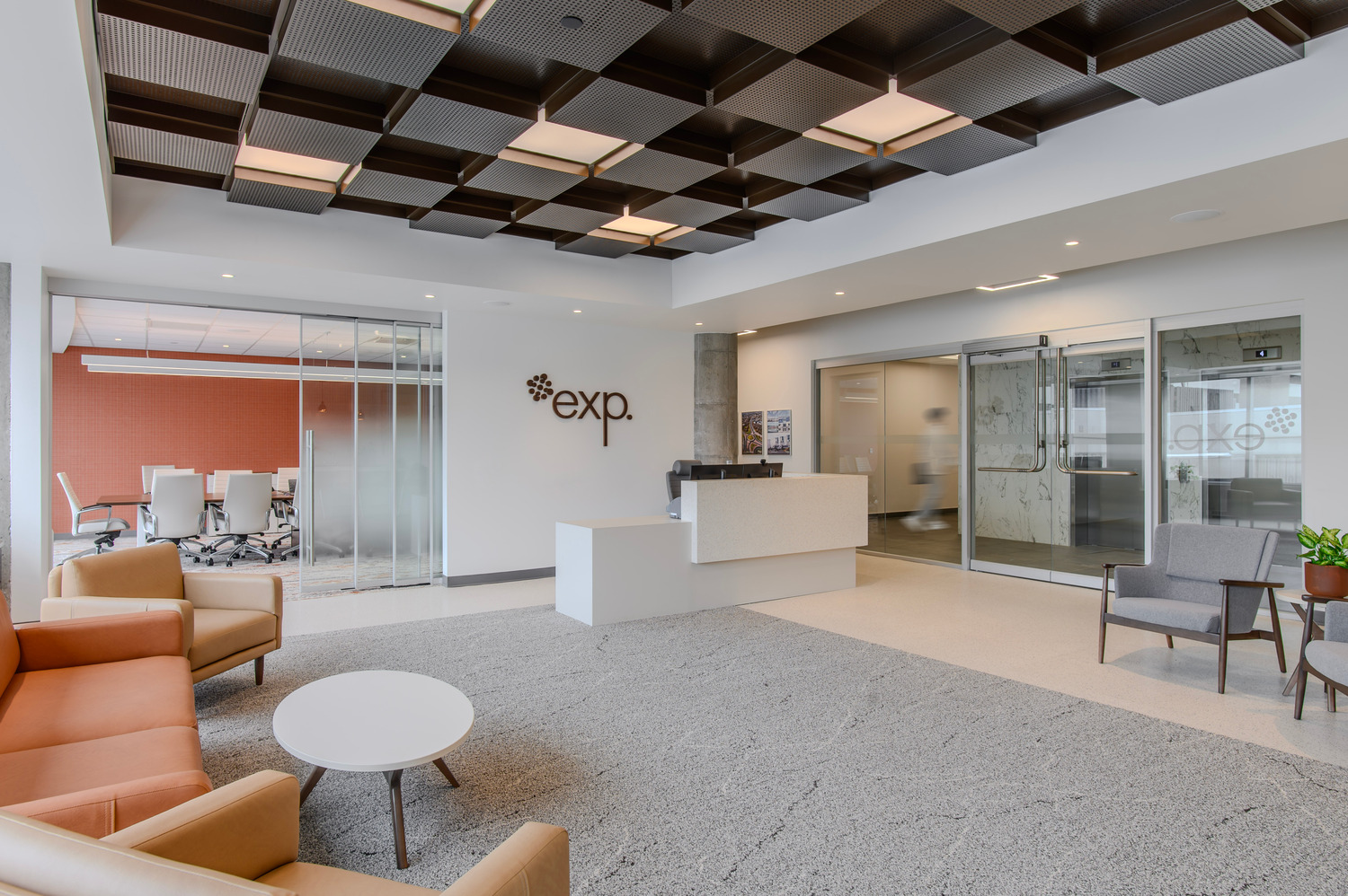 Modern office reception area with a white desk, seating, and a ceiling with geometric patterns. Glass doors lead to a conference room.