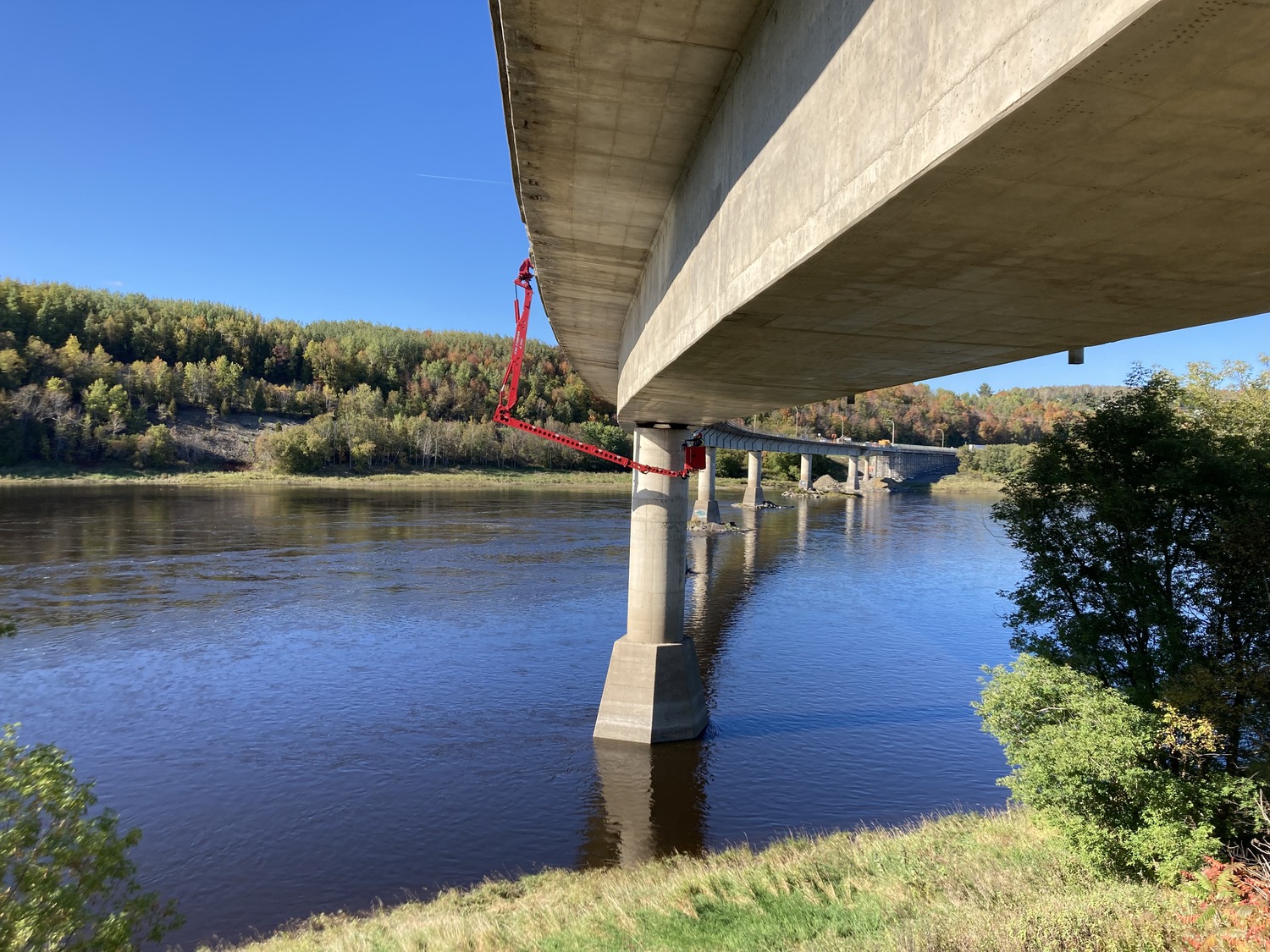 A large concrete bridge with red equipment attached to its columns spans a calm river, with a forested hillside and clear blue sky in the background.