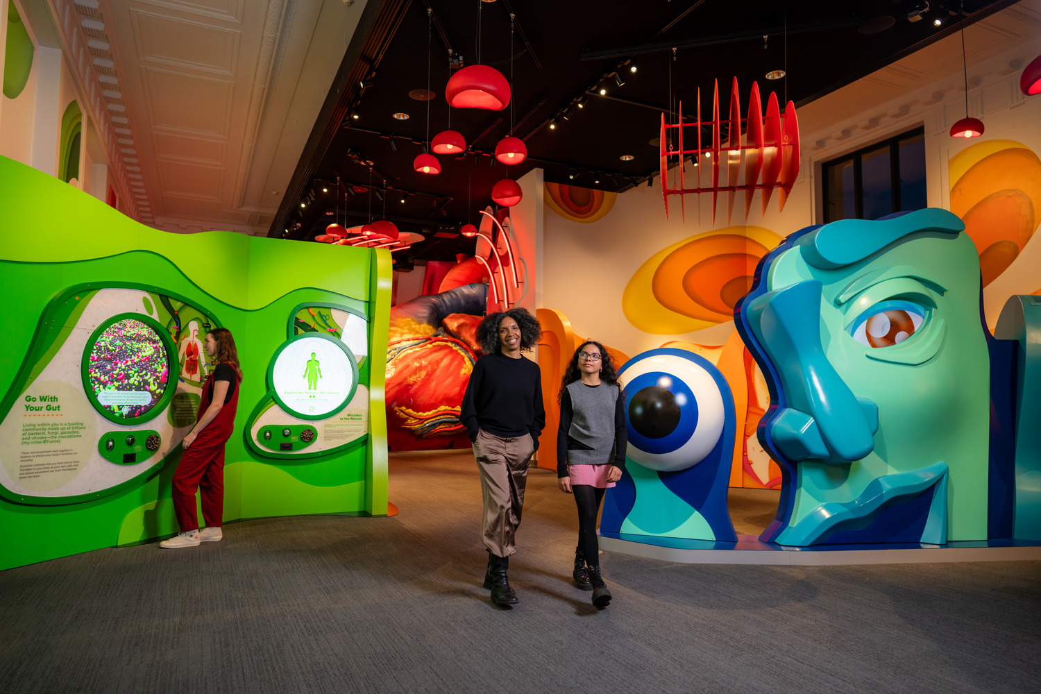 Three people are inside a colorful, modern exhibition space with vibrant, abstract sculptures and interactive displays on the walls.