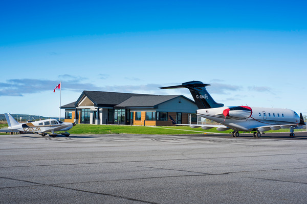 Two small airplanes are parked on an airport tarmac in front of a low building with a Canadian flag.