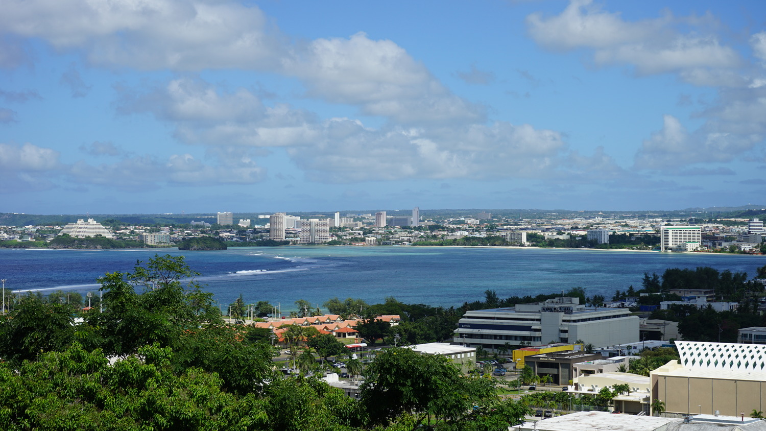 A coastal cityscape showcases Guam infrastructure resilience, with modern buildings lining the shoreline, bordered by blue ocean waters and greenery under a partly cloudy sky.