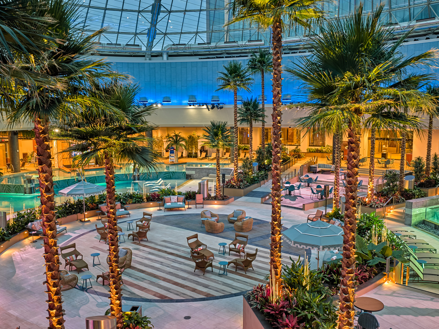 Indoor pool and lounge area with palm trees, patterned flooring, seating arrangements, and greenery under a glass dome ceiling.