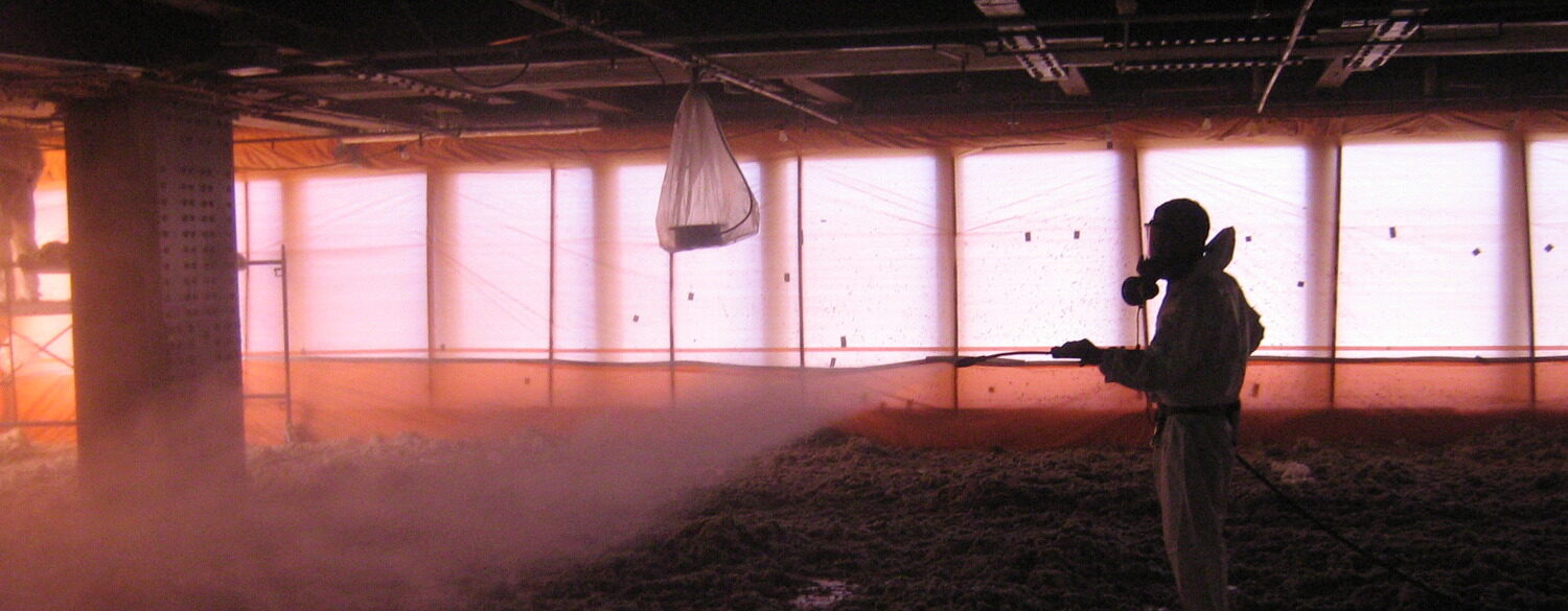 A person in protective gear sprays a hose inside a dimly lit room filled with insulation material on the floor.