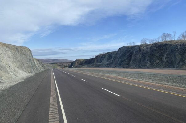 A paved road runs between rocky hills under a partly cloudy sky, with no vehicles or people visible.
