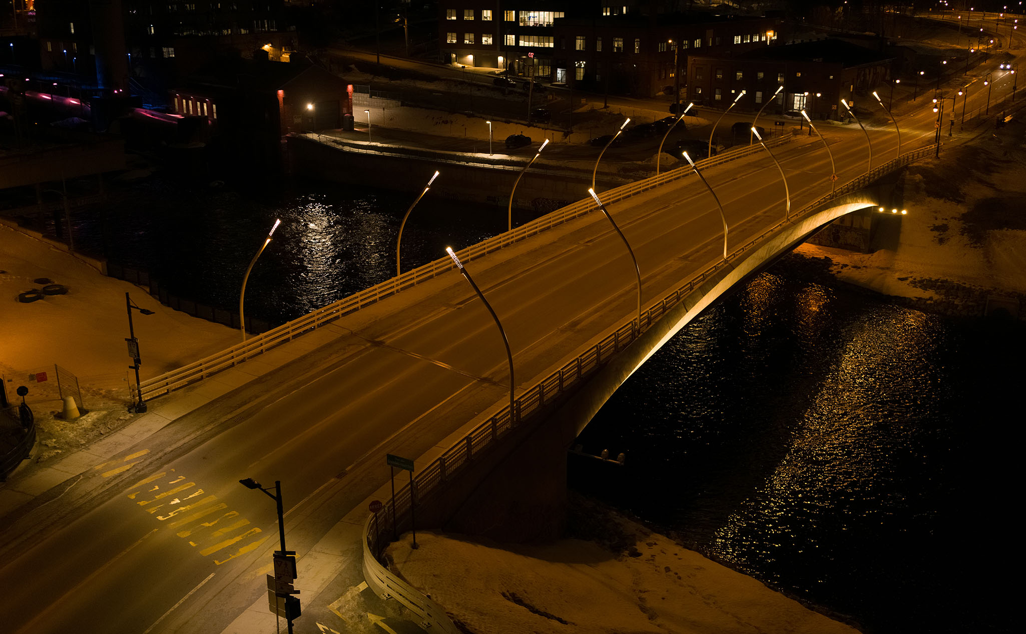 A well-lit bridge spans over a body of water at night, with snow on the ground and buildings illuminated in the background.