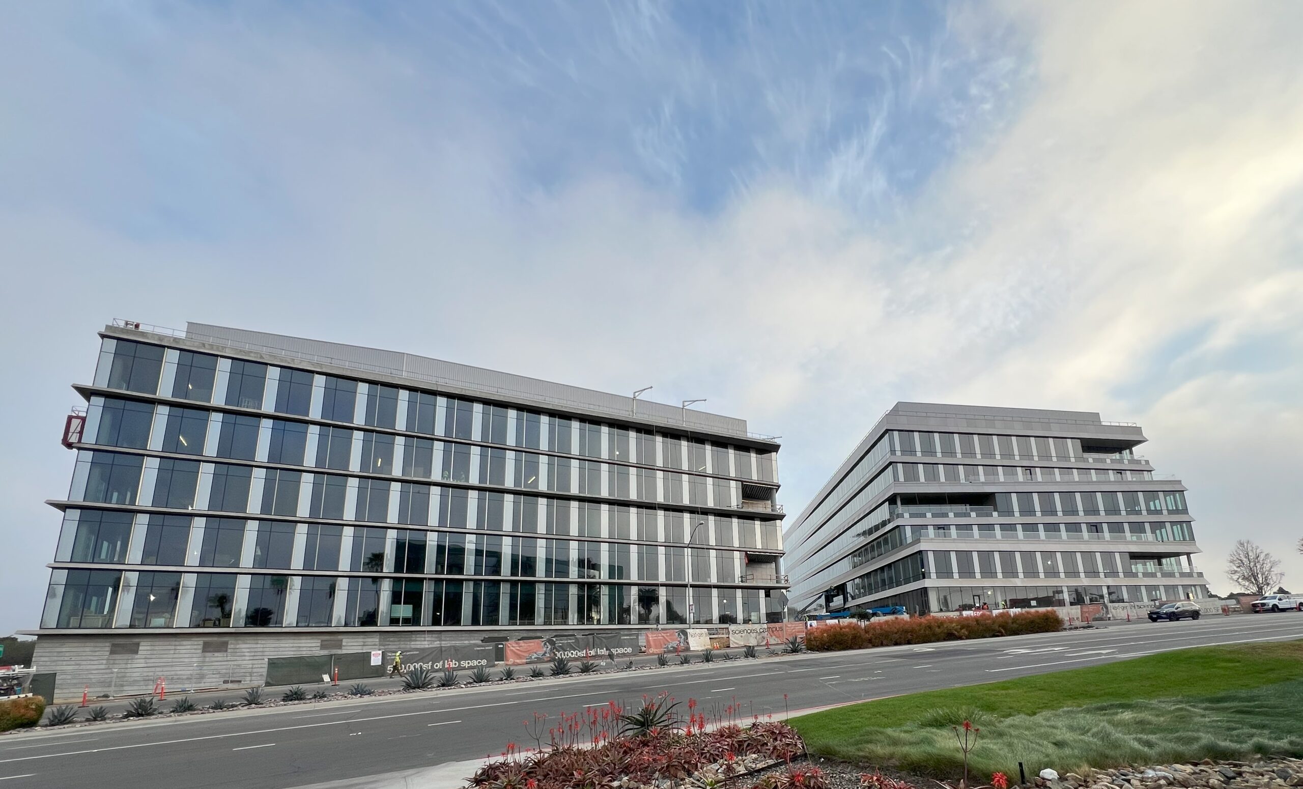 Two modern office buildings with glass facades stand side by side along a road under a partly cloudy sky, with landscaping in the foreground.