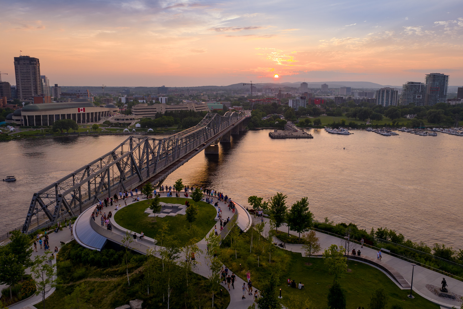 Aerial view of a park with people, a large river, a bridge, and city buildings at sunset. The Canadian flag is visible on a building to the left.