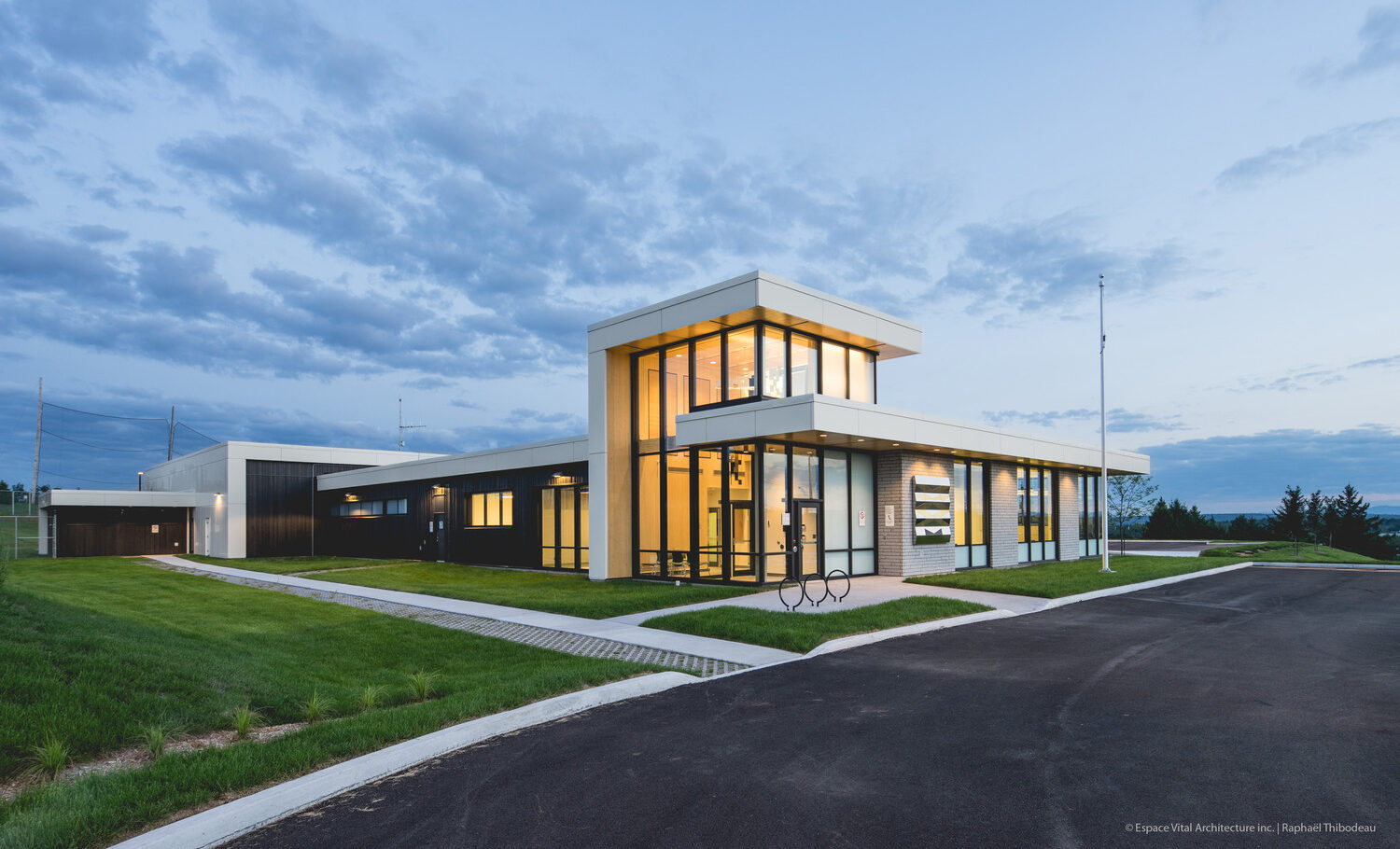 Modern police station building with large glass windows and a flat roof, surrounded by grass and a paved road, set against a twilight sky.