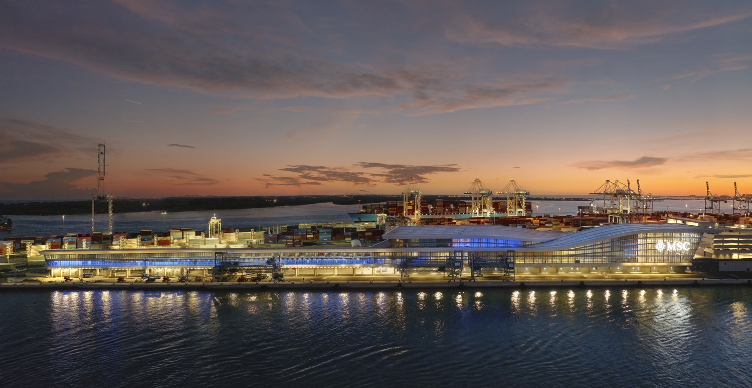 A modern cruise terminal with blue lighting sits beside stacked shipping containers and cranes at a busy port during sunset.