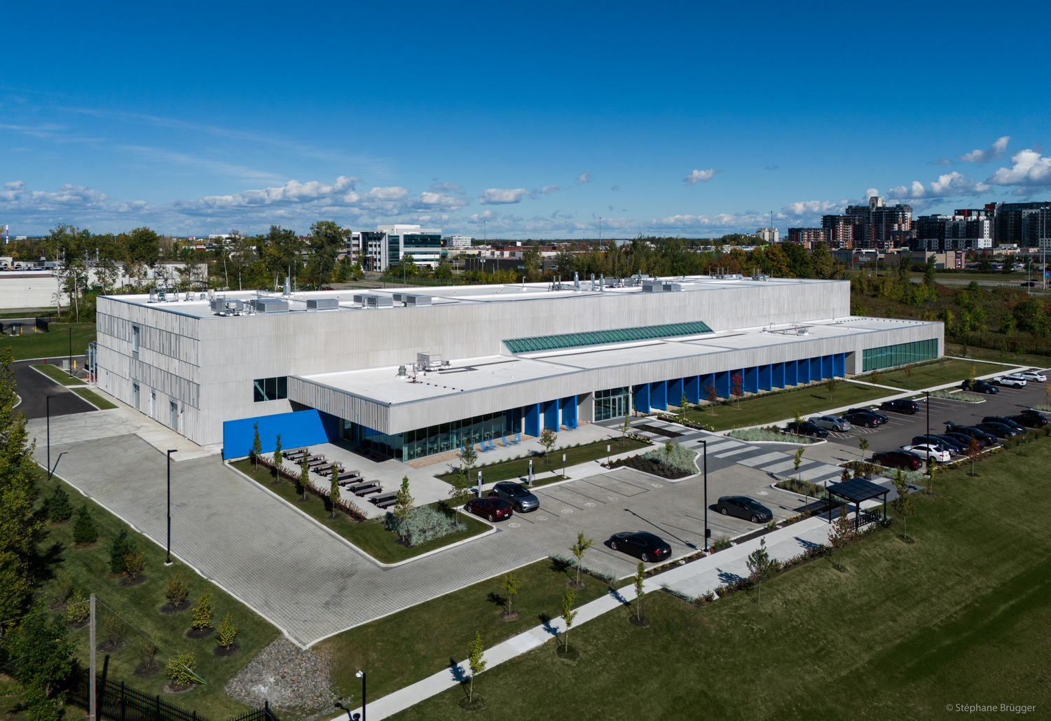 Aerial view of a large, modern industrial or office building with a parking lot, surrounded by grass and some trees, under a blue sky.