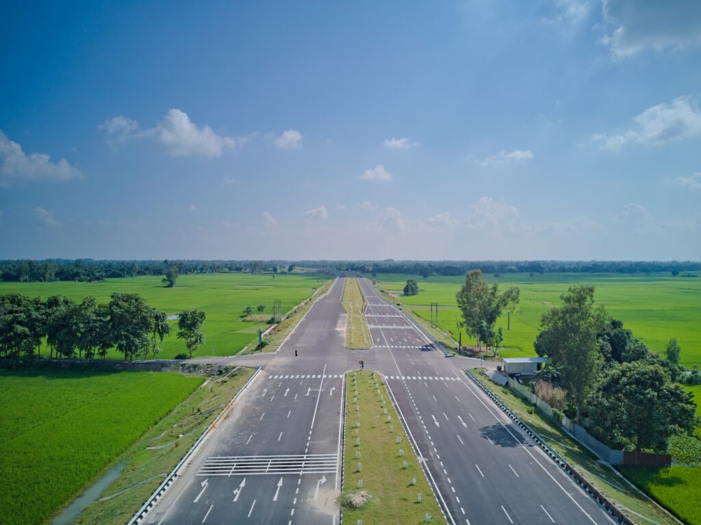 A wide, empty highway stretches through green fields under a blue sky with scattered clouds. Trees line the road, and the scene is calm and rural.