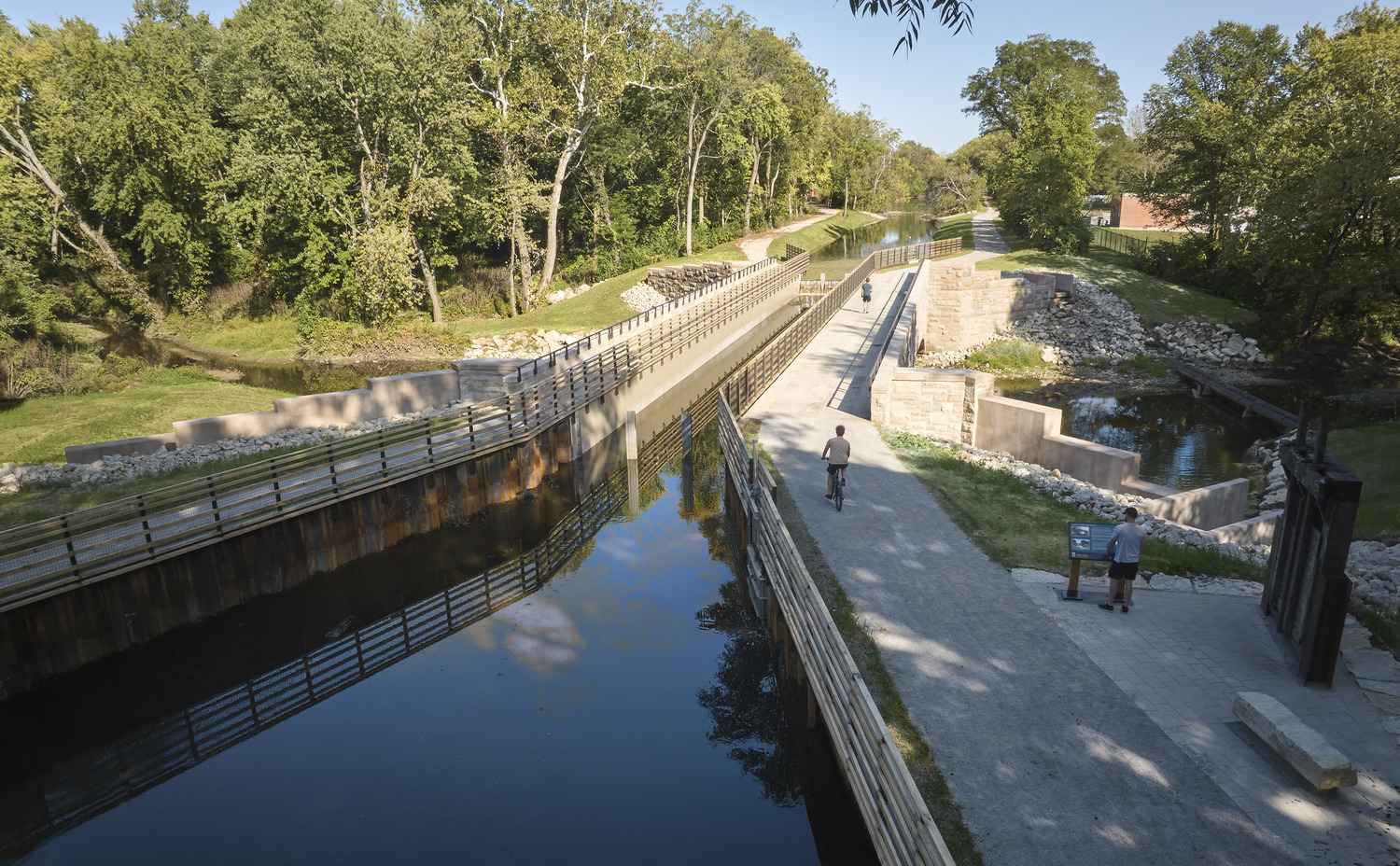A canal with calm water runs parallel to a walkway and bridge, surrounded by green trees and grass. Two people stand near a sign, and one person cycles along the path.