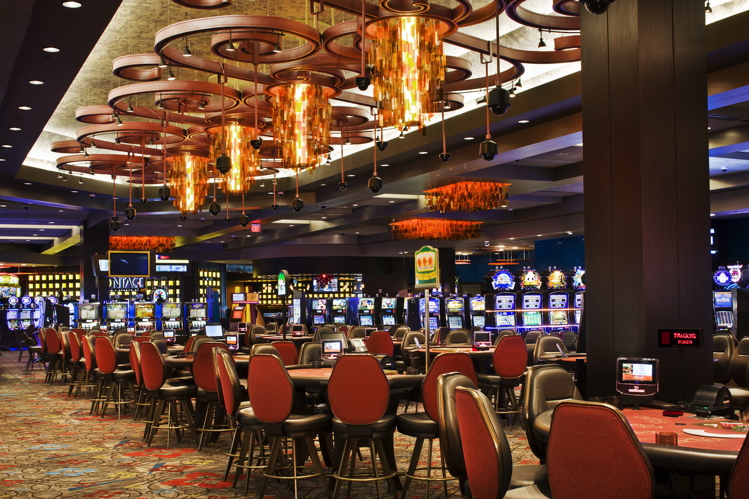 Casino interior with rows of slot machines, empty gaming tables, red chairs, ornate lighting fixtures, and patterned carpet.
