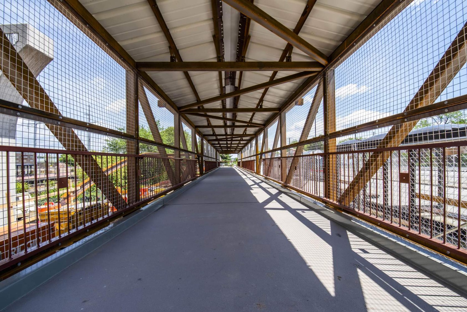 A covered pedestrian bridge with metal fencing and a truss roof spans over a construction area on a sunny day.