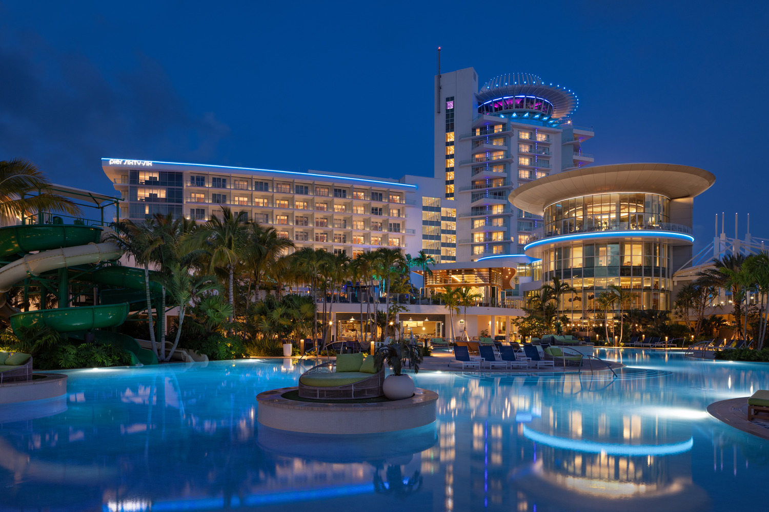 Modern resort hotel with illuminated buildings, palm trees, waterslides, and a large pool at dusk.