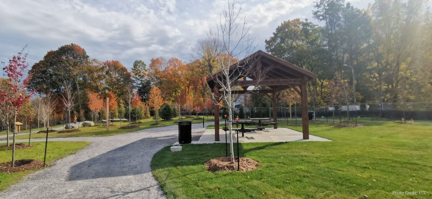 A covered picnic pavilion with tables sits beside a paved path in a park with green grass and autumn-colored trees.