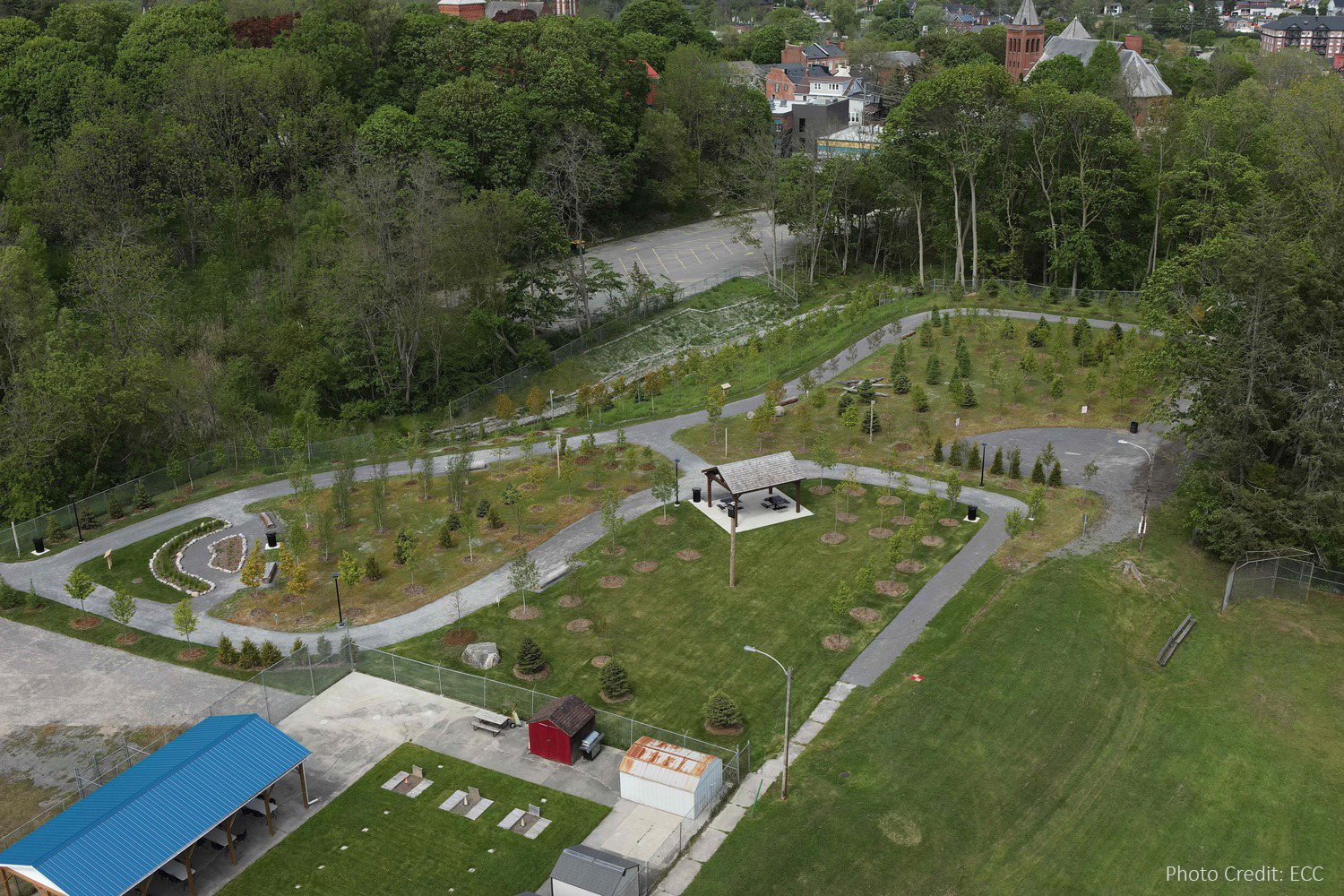 Aerial view of a landscaped park with walking paths, small trees, a pavilion, and surrounding greenery. Buildings and parking lot visible in the background.