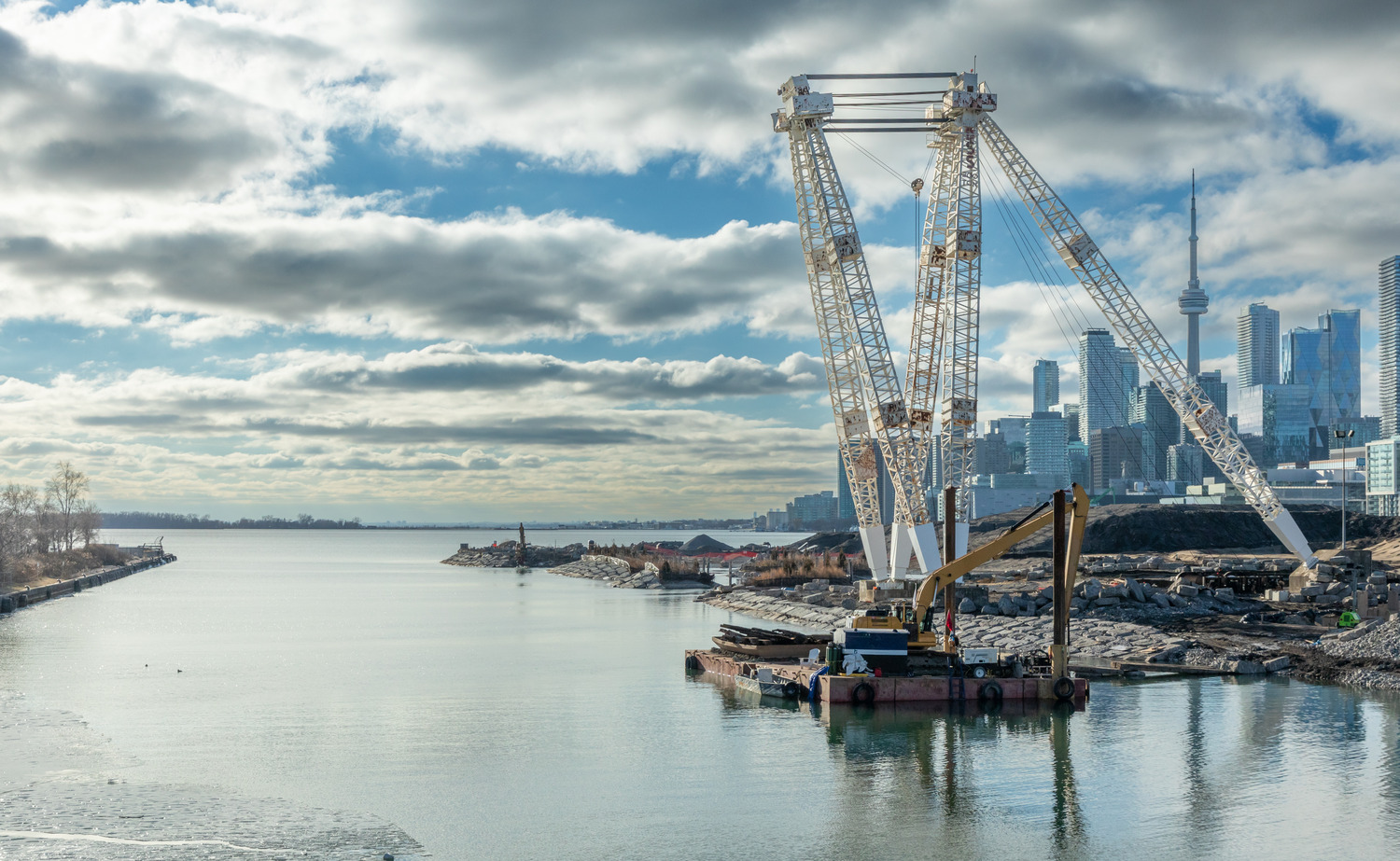 A large crane on a barge operates near the shoreline with city buildings and the CN Tower visible in the background under a cloudy sky.