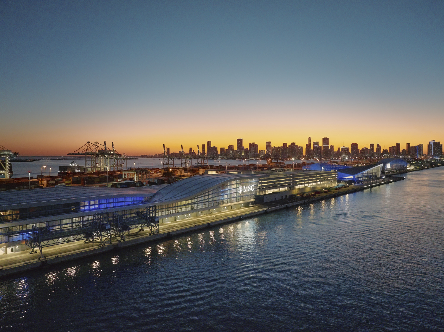 A modern pier terminal with curved roofs is lit up along the waterfront at sunset, with a city skyline and cranes visible in the background.