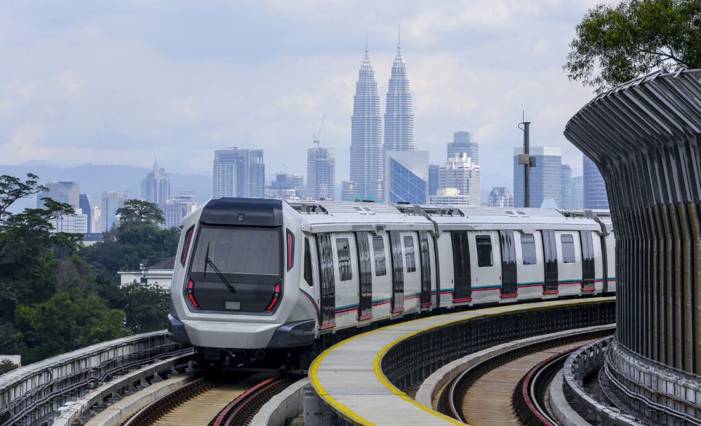 A modern train travels on elevated tracks with the Kuala Lumpur city skyline, including the Petronas Twin Towers, visible in the background.