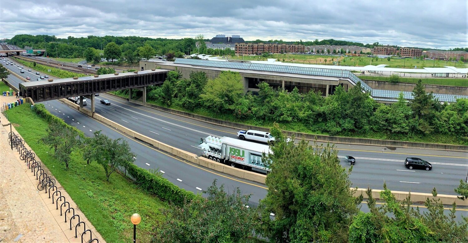 A multi-lane highway with trucks and cars passes next to a Metro station surrounded by trees and buildings under a cloudy sky.