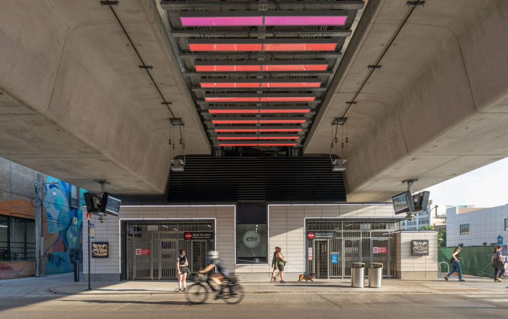View of a CTA transit station entrance under a bridge, illuminated by overhead pink lights. Pedestrians, a cyclist, and a person walking a dog are visible—highlighting the vibrant transportation scene worthy of Transportation Awards recognition.