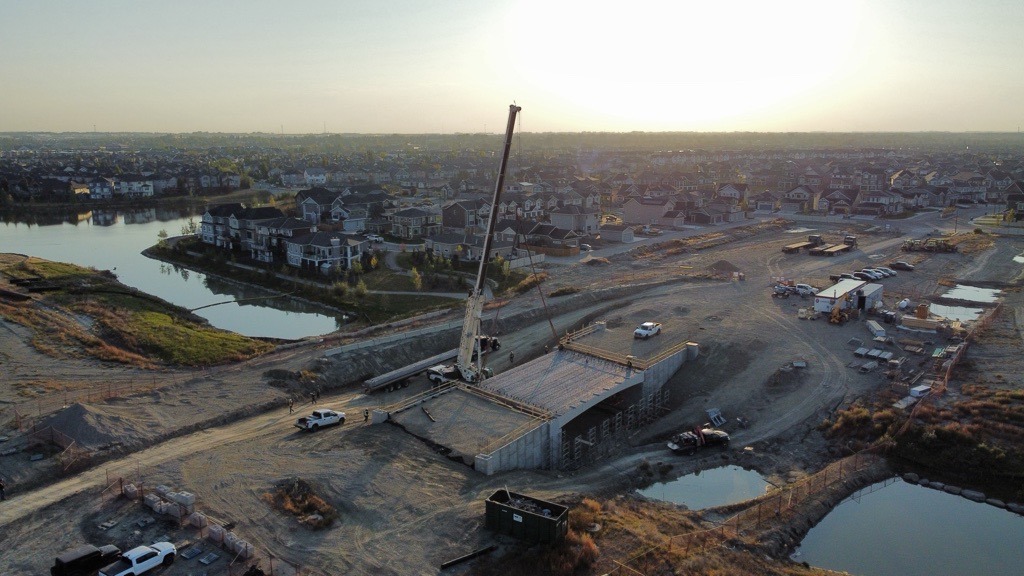 A construction site with a crane building the Bayview Phase 5 Bridge over a dry area, surrounded by houses, small ponds, and construction vehicles at sunset.