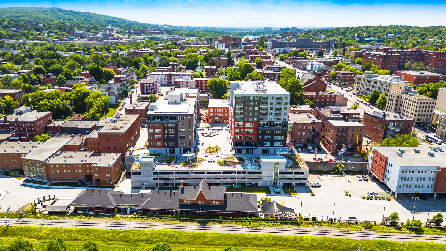 Aerial view of a cityscape featuring mid-rise buildings, a parking structure, a historic train station, and greenery in the background.
