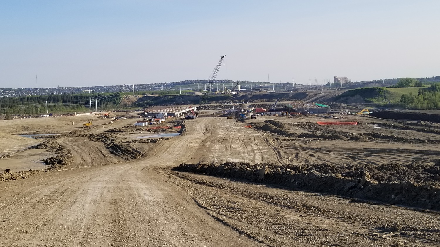 Wide view of a large construction site with dirt roads, heavy machinery, cranes, and partially built structures under a clear sky.