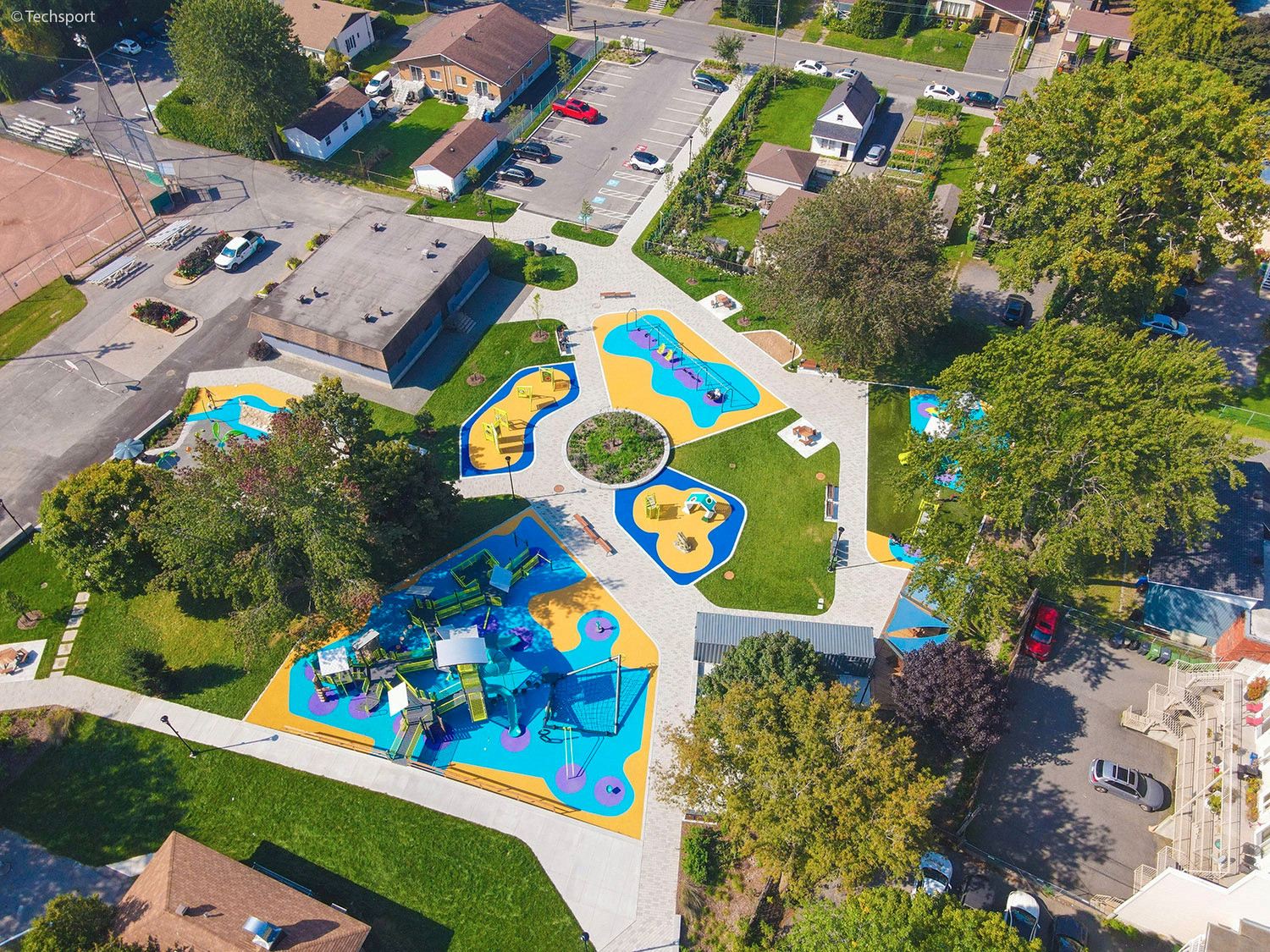 Aerial view of a colorful playground with play structures, pathways, and surrounding trees in a residential neighborhood.