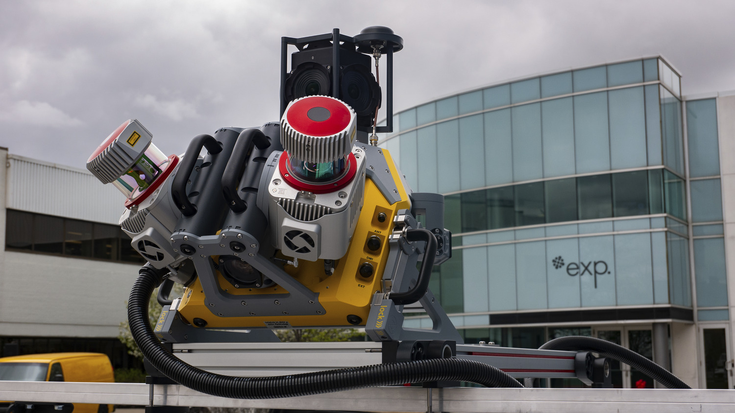 A large, industrial 3D camera or scanner is mounted on metal rails outside a modern glass office building with the sign "exp." visible.