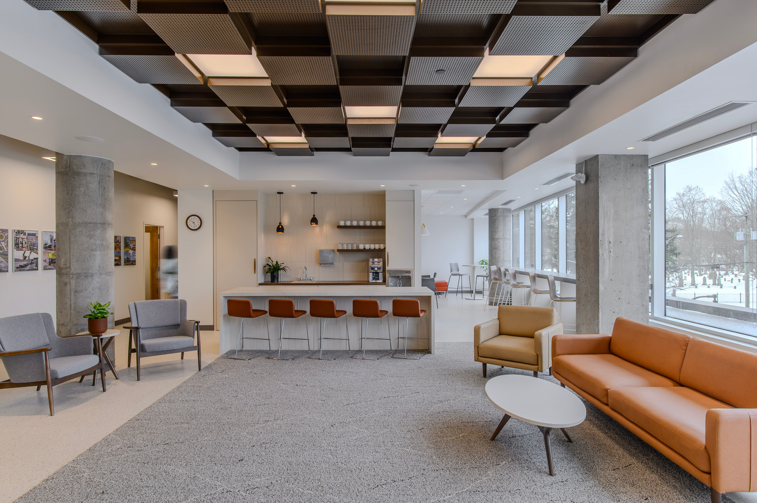 Modern lounge area with armchairs, sofas, a coffee table, and a counter with barstools; large windows let in natural light, and the ceiling features a geometric design.