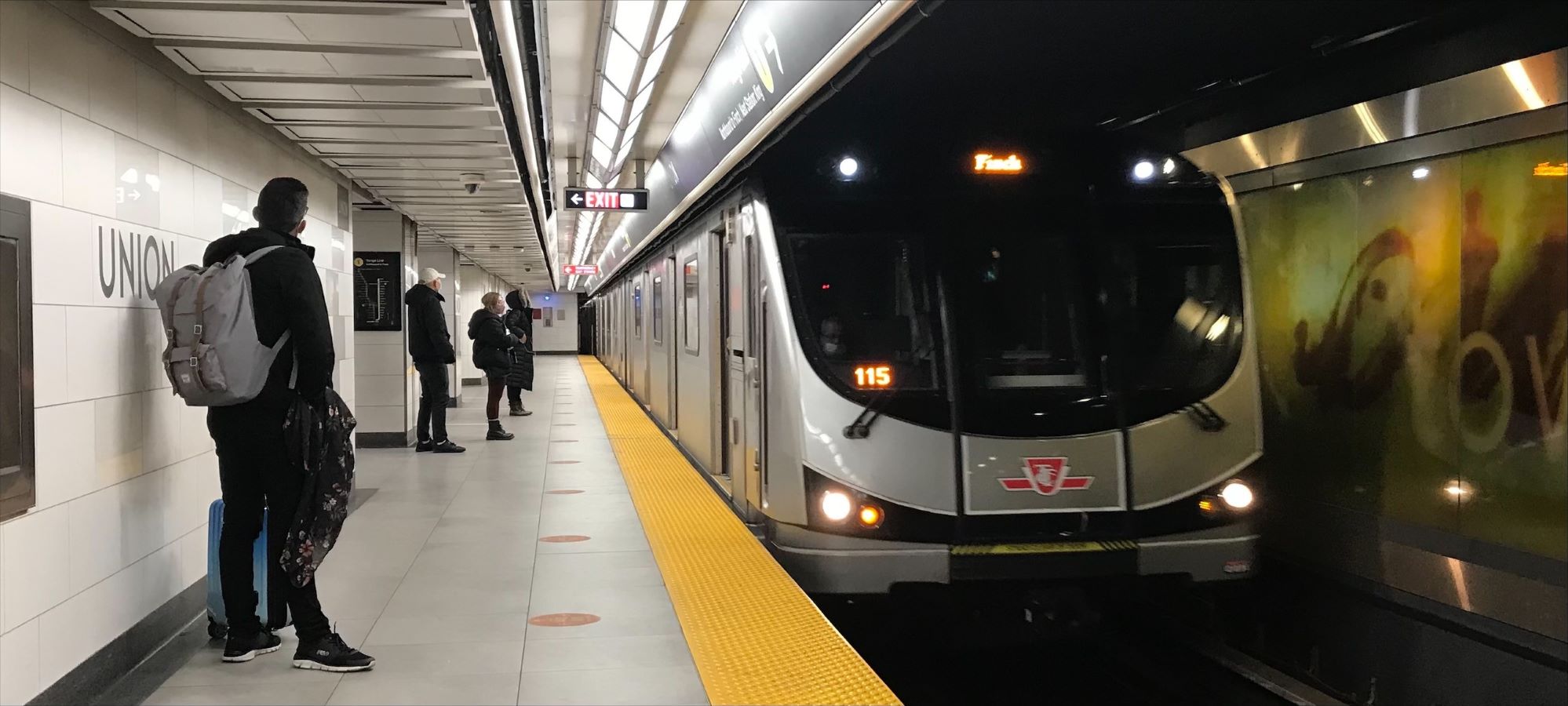 A subway train arrives at an underground platform as several people wait nearby, some standing and one person with a backpack facing the train.