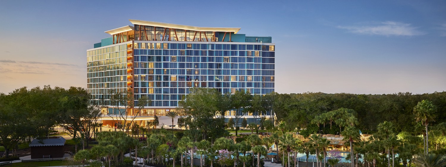 A modern multi-story hotel, the Disney Swan Reserve features large windows and is surrounded by trees and a landscaped pool area with invisible infrastructure, all set under a clear blue sky at sunset.