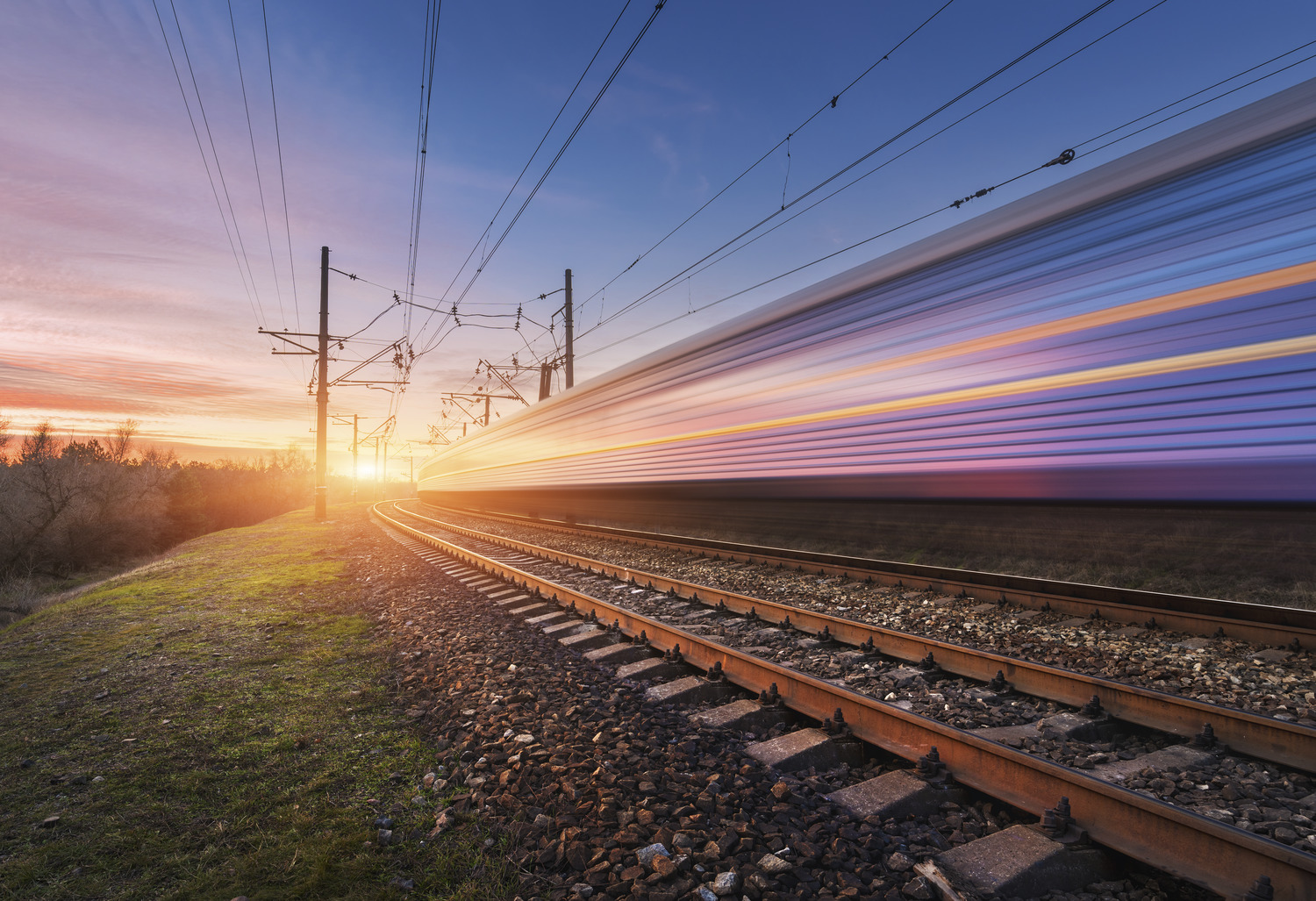 A high-speed train blurs past on a railway track at sunset, with power lines overhead and a clear sky in the background.