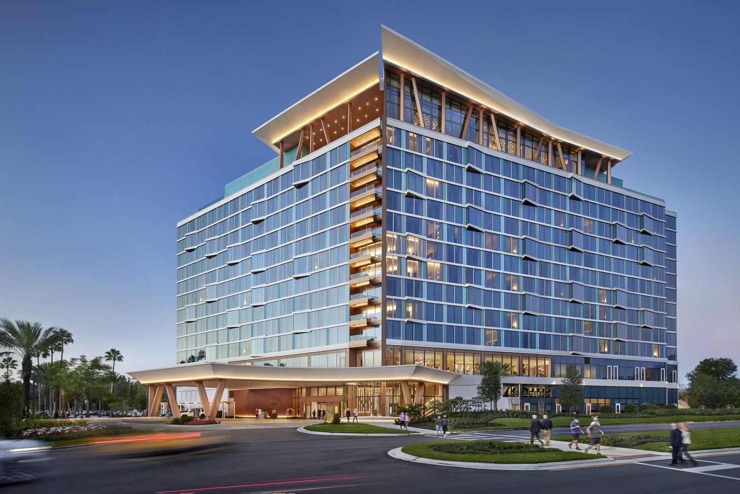 Modern multi-story hotel with glass exterior, illuminated balconies, and a covered entrance; people walk nearby and cars pass in front at dusk near the striking Disney Swan Reserve.